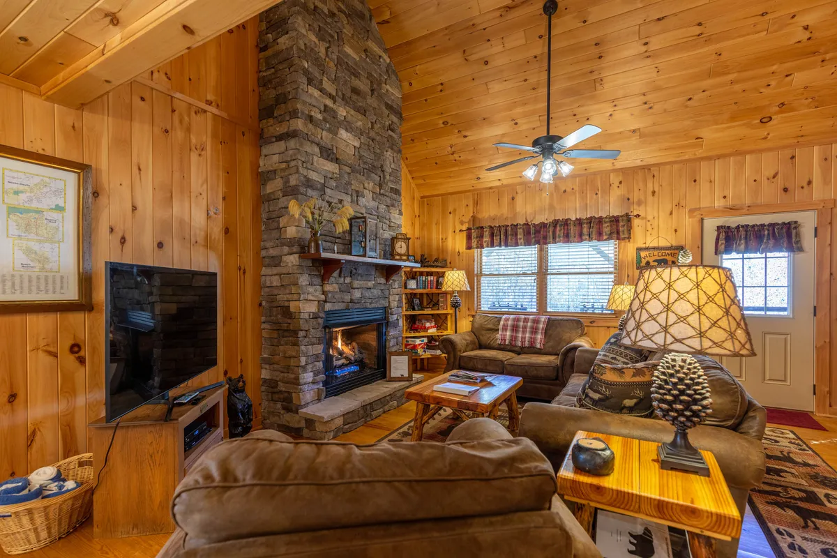 A warm, inviting living room set inside a rustic log cabin, featuring a grand stone fireplace, plush seating, exposed wooden beams, and soft ambient lighting. The space is arranged for comfort and gathering, perfect for cozying up after a day in the mountains.
