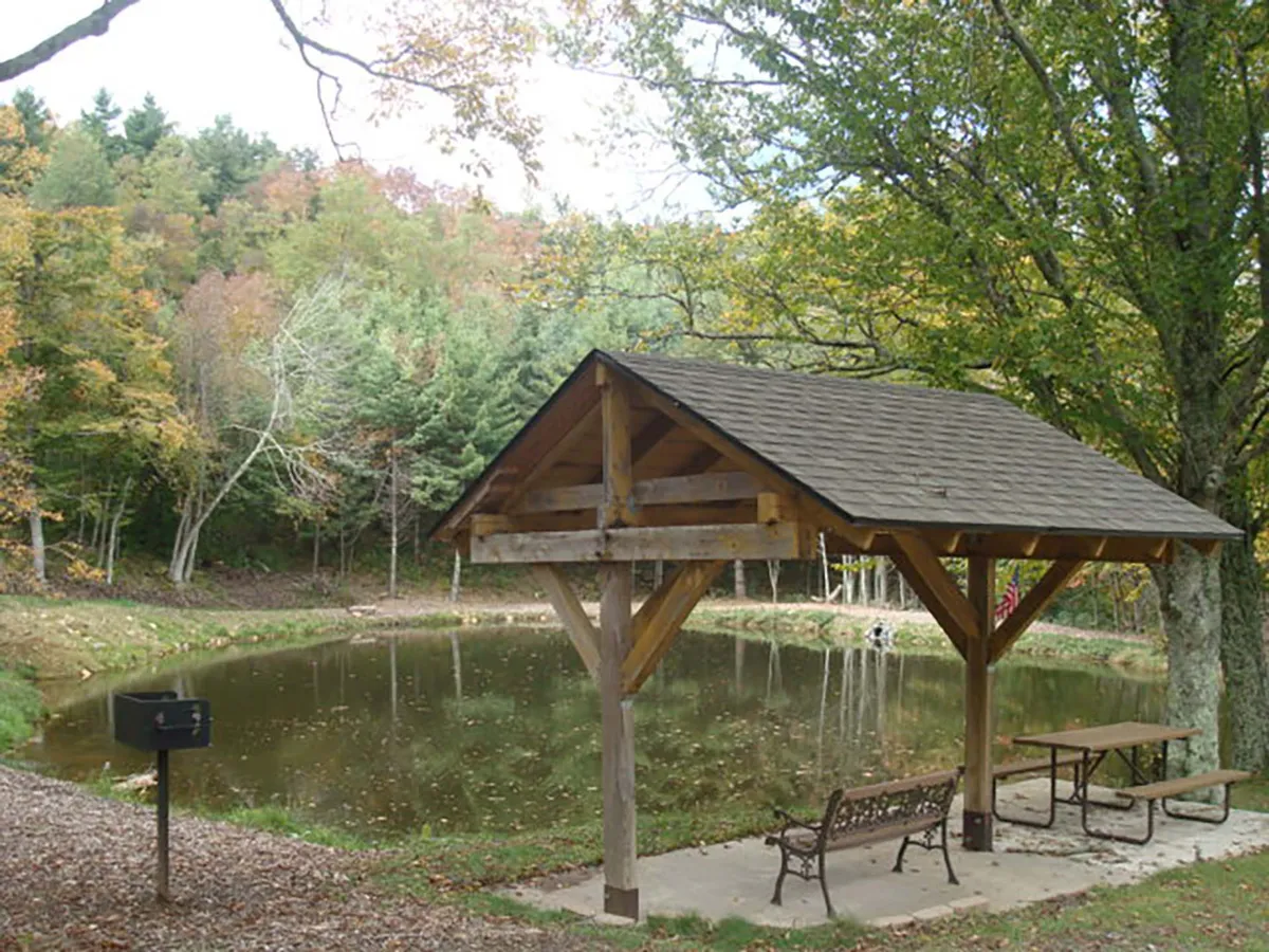 This image features a serene pavilion set beside a pond, surrounded by mature trees and a bench. A grill is visible, suggesting spaces for picnicking and outdoor gatherings. The tranquil water reflects the surrounding nature, setting a relaxing, family-friendly atmosphere.