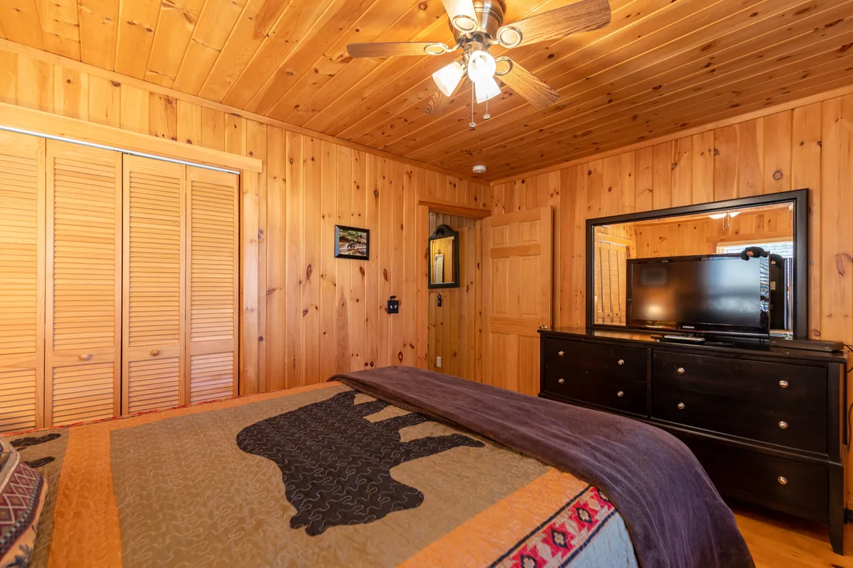 A bedroom with wood paneling, a bed covered in a patterned quilt, a large mirror, and a dresser with a television. Warm, natural light fills the room, creating a cozy spot for relaxing or enjoying TV in bed.