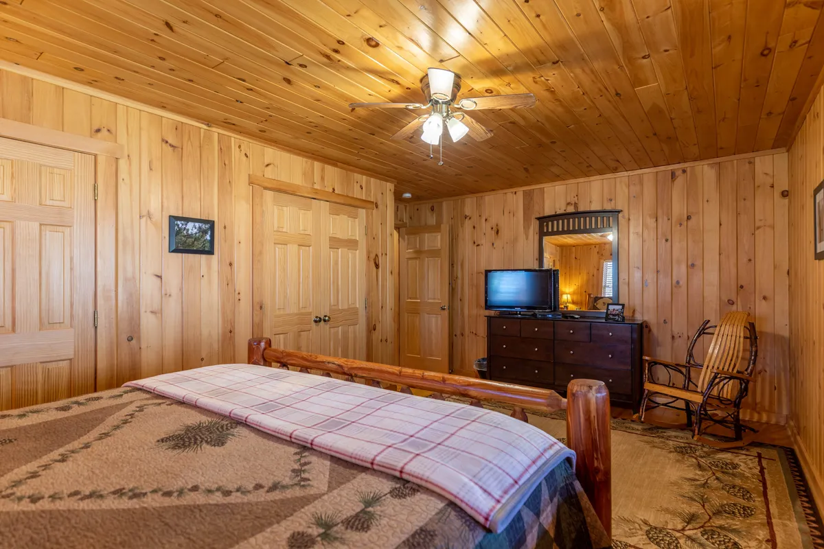 A spacious bedroom with a wooden bed frame, ceiling fan, and a television across from the bed. The room also has a cozy rocking chair and a decorative mirror, creating a space for both relaxation and private entertainment.