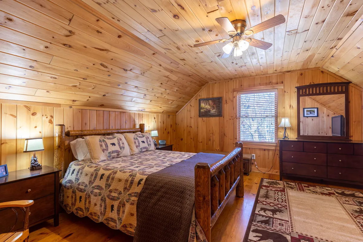 This image features a warmly lit bedroom with a wooden bed, plaid bedding, wood-paneled ceiling, and nightstands with lamps on each side. A rug adds to the cozy atmosphere. Sunlight streams in, highlighting the rustic charm and inviting feel of the space.