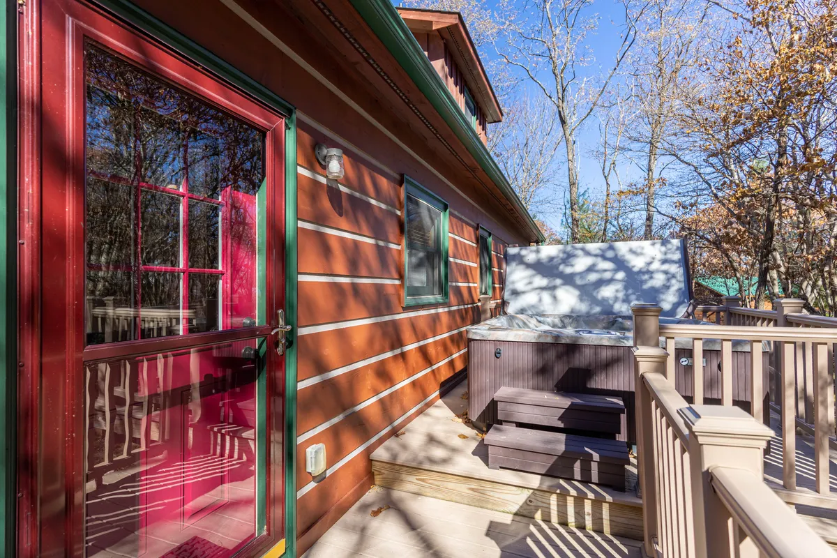 This exterior shot features a private wooden deck nestled among trees, with a red door leading to the cabin and a secluded hot tub as the focal point. The hot tub is surrounded by natural scenery, providing a tranquil space for relaxation and connection with nature.