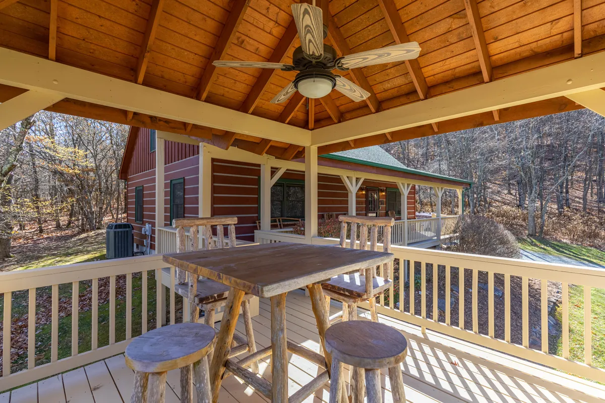 This exterior image captures a spacious outdoor dining area with a wooden table and bar stools beneath a ceiling fan and exposed wooden beams. The backdrop features the house and lush, green surroundings, evoking a sense of openness and connection to nature.